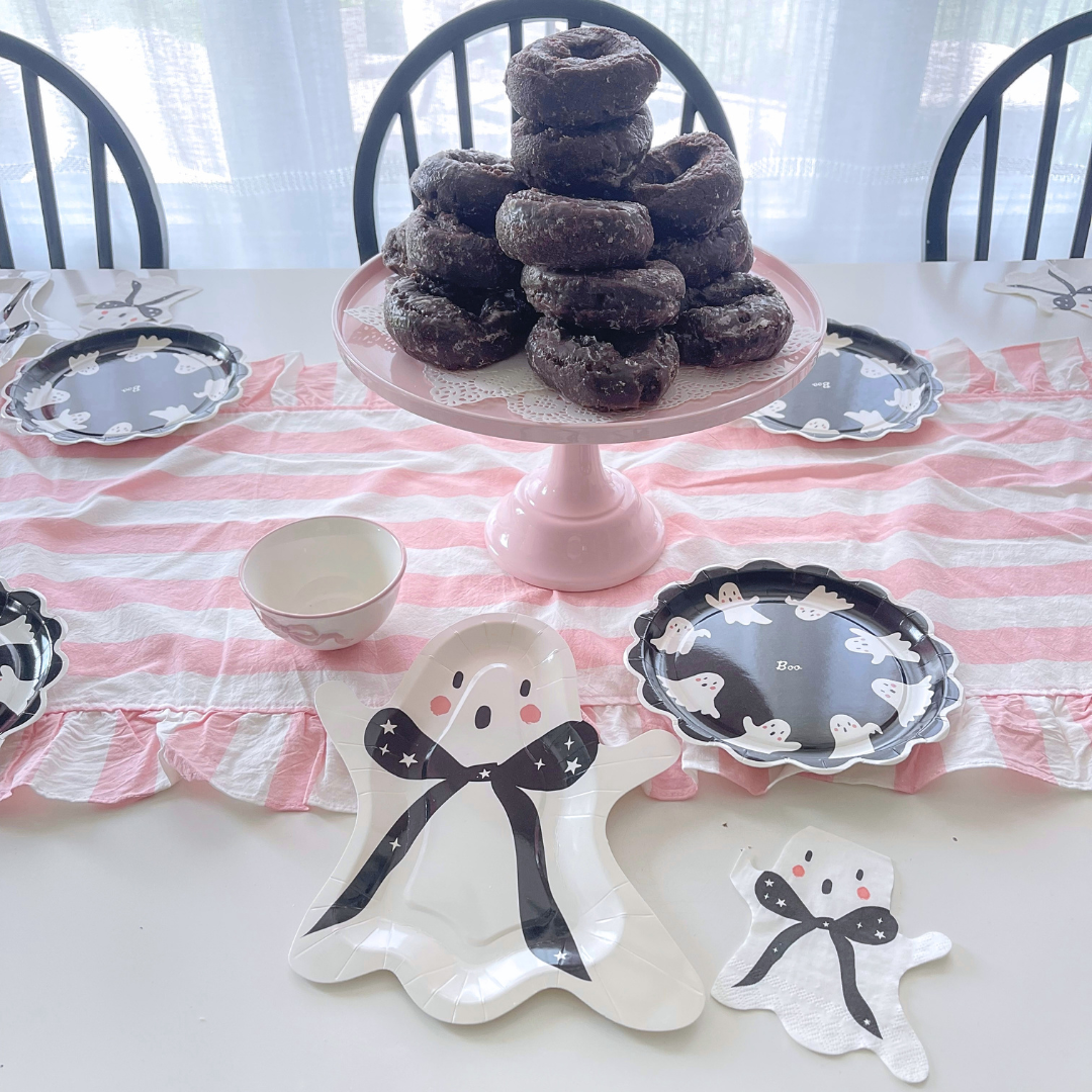 A styled dining table setting featuring a pink and white striped table runner, black and white ghost-shaped plates, a pink cake stand holding a stack of chocolate-covered donuts, and a small white bowl.