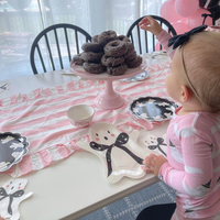 A baby in a pink pajama shirt with a black bow headband stands at a table decorated for a party. The table is covered with a pink and white striped runner, matching plates, and a pink cake stand holding chocolate donuts.