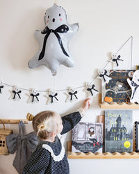Close view of the double-sided ghost mylar balloon with black bow and pink cheeks, a child reaching toward a ghost garland below