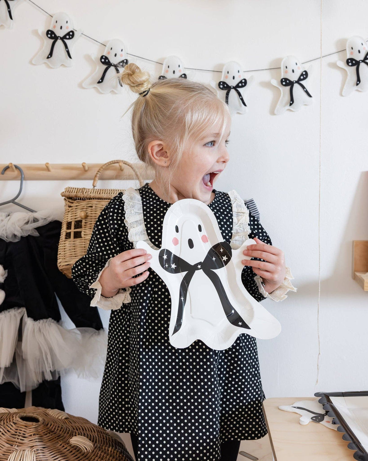 Child holding the ghost-shaped paper plate with a bow tie, standing in a room with Halloween decorations.