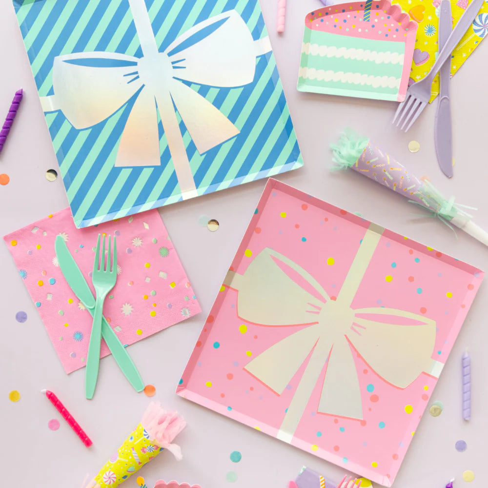 Overhead party scene showing pink and blue gift-bow plates mixed with confetti napkins, cake-slice plates, and noisemakers on a pastel table.