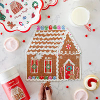 Hand placing the last piece in a gingerbread house jigsaw puzzle beside its red tube; plates and cookies shown separately, not included
