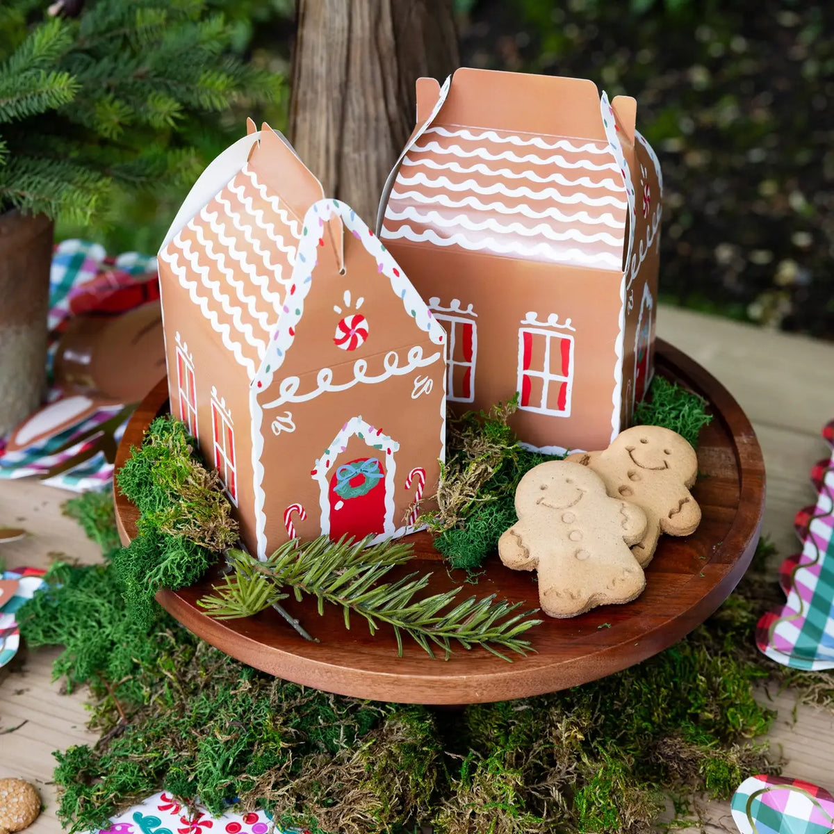 2 gingerbread house treat boxes on a wooden cake stand with gingerbread cookies and moss