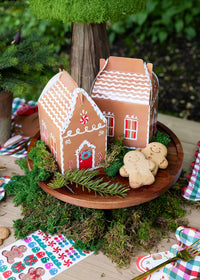 2 gingerbread house treat boxes on a wooden cake stand with gingerbread cookies and moss
