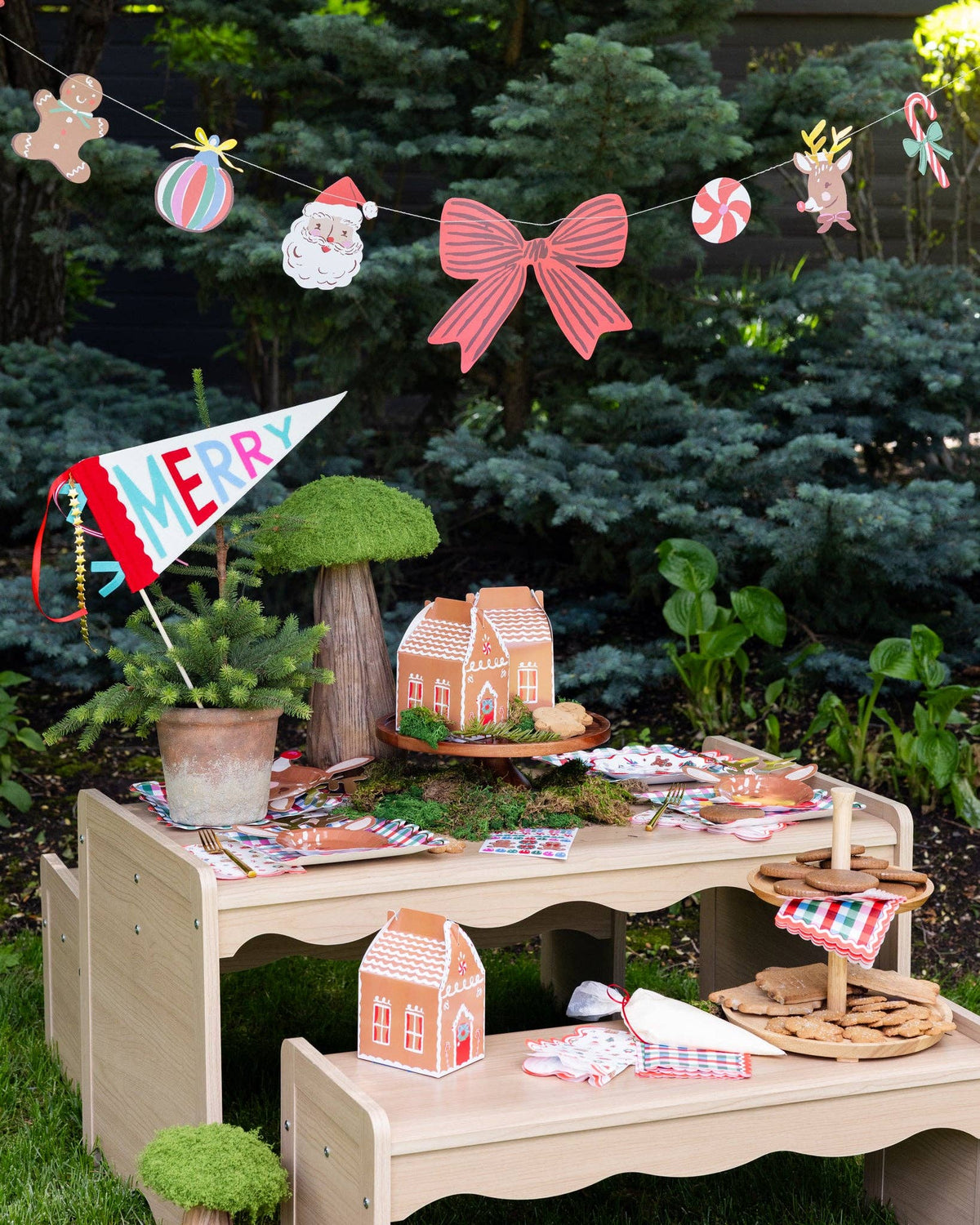 Gingerbread house treat boxes on an outdoor kids' party table under a Christmas icon garland; MERRY pennant, plates, and napkins shown separately, not included