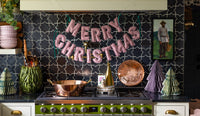 Red gingham merry christmas banner hanging over kitchen stove with copper pots and paper trees