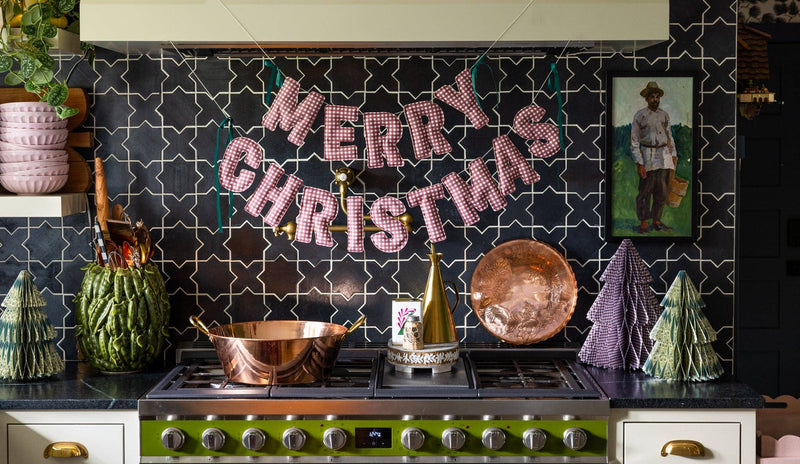 Red gingham merry christmas banner hanging over kitchen stove with copper pots and paper trees