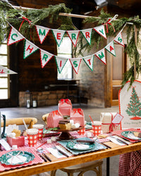 Rustic table with red gingham treat boxes, plaid tableware, a tree plate, and a "Merry Christmas" felt banner.