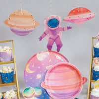 Pink space party dessert table with a hanging girl astronaut, pastel planet cutouts, a spaceship, watercolor planet plates, and popcorn cups.