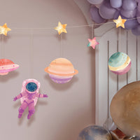 Pink and purple watercolor space banner with a girl astronaut, pastel planets, gold-foil stars, and a spaceship, strung above a purple balloon garland.