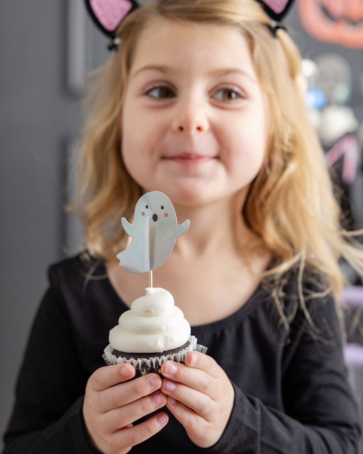 Young girl in a black shirt smiling while holding a frosted cupcake decorated with a die-cut paper ghost topper.