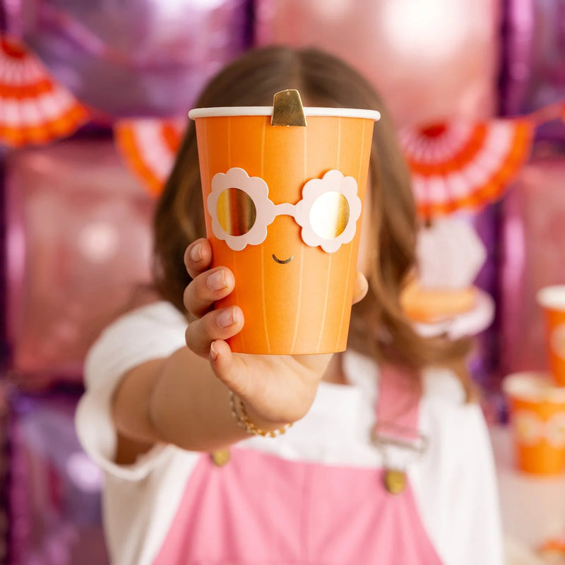 Child in pink overalls holds one Groovy Pumpkin paper cup toward camera, showing smiling face and shiny sunglasses for playful Halloween vibe.