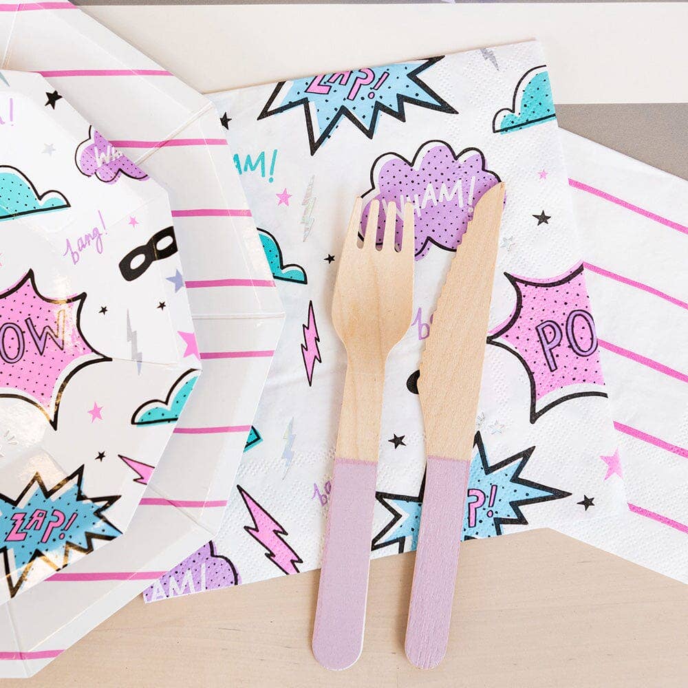 Flat lay of a girl power party table featuring large superhero napkins, octagonal plates, and purple wooden cutlery.