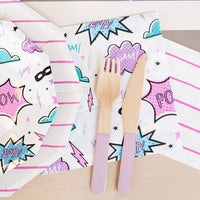 Flat lay of a girl power party table featuring large superhero napkins, octagonal plates, and purple wooden cutlery.
