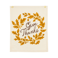 Overhead studio view of a rectangular off-white canvas banner featuring a mustard yellow oak leaf wreath and black Give Thanks text.