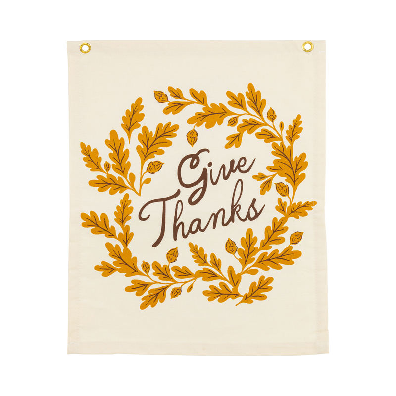 Overhead studio view of a rectangular off-white canvas banner featuring a mustard yellow oak leaf wreath and black Give Thanks text.