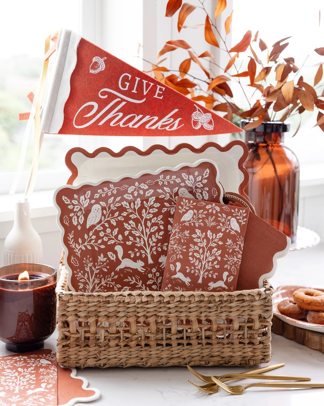 Fall mantel decor styling — the Give Thanks pennant in a woven basket with rust folk-art napkins, a lit candle, and a bottle of branches on a cozy autumn surface.