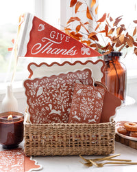 Fall mantel decor styling — the Give Thanks pennant in a woven basket with rust folk-art napkins, a lit candle, and a bottle of branches on a cozy autumn surface.