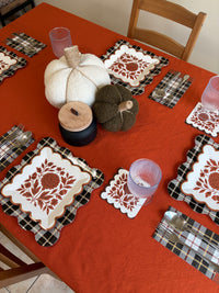 Thanksgiving table setting with rust floral square paper plates, white and green knit pumpkins, a black candle with a wood lid, pink glasses, and black and white plaid placemats on an orange tablecloth