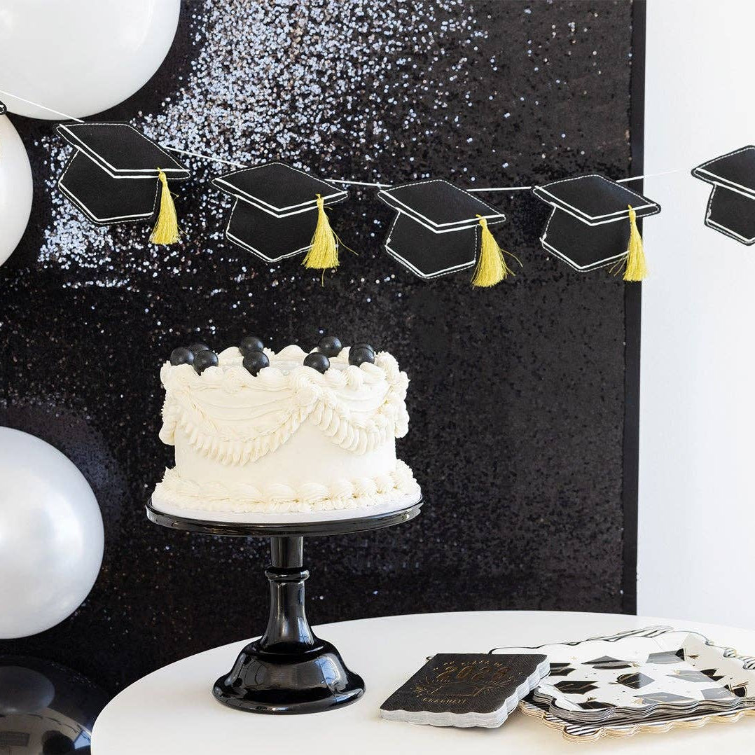 A black felt graduation cap garland hung above a white frosted cake, showing a matte, photo-ready finish.