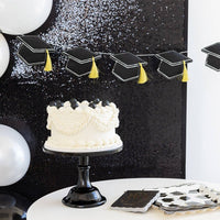 A black felt graduation cap garland hung above a white frosted cake, showing a matte, photo-ready finish.
