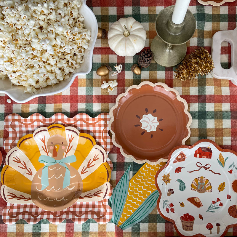 Turkey plate on a gingham plate next to a pie plate, corn napkin and harvest plate on a fall plaid tablecloth with popcorn and a white pumpkin
