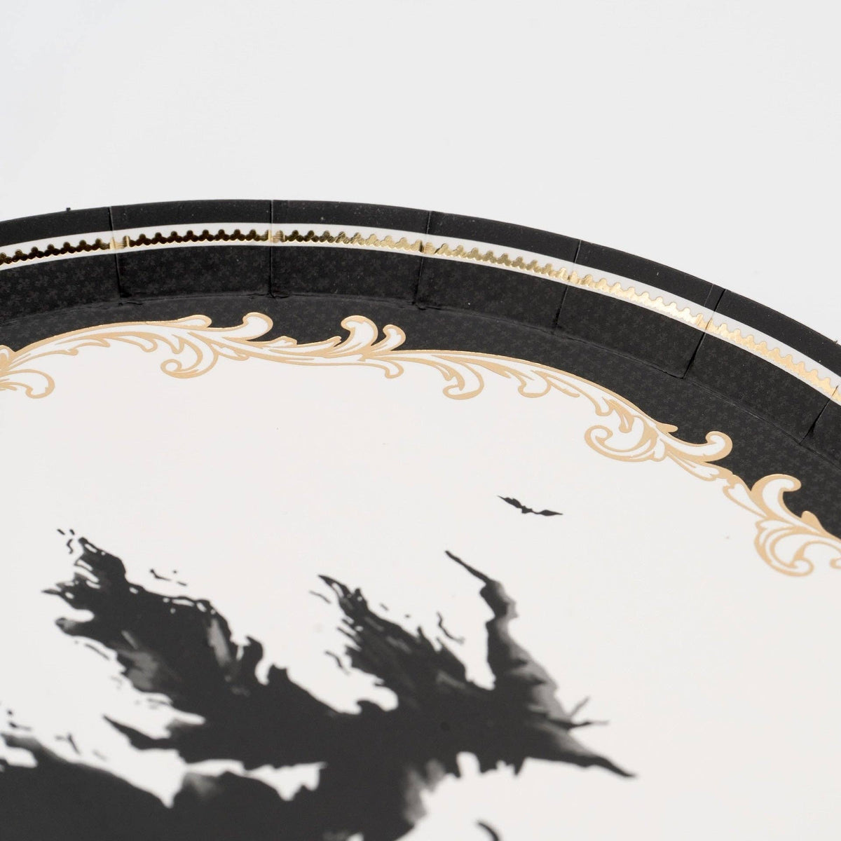 Macro detail shot highlighting the metallic gold foil filigree border and black flying witch graphic on the white paper dessert plate.
