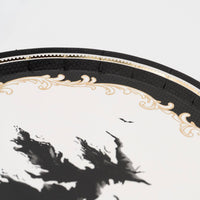Macro detail shot highlighting the metallic gold foil filigree border and black flying witch graphic on the white paper dessert plate.