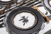 Angled place setting view showing a gold fork resting beside a vintage witch plate and black charger on a striped paper runner.