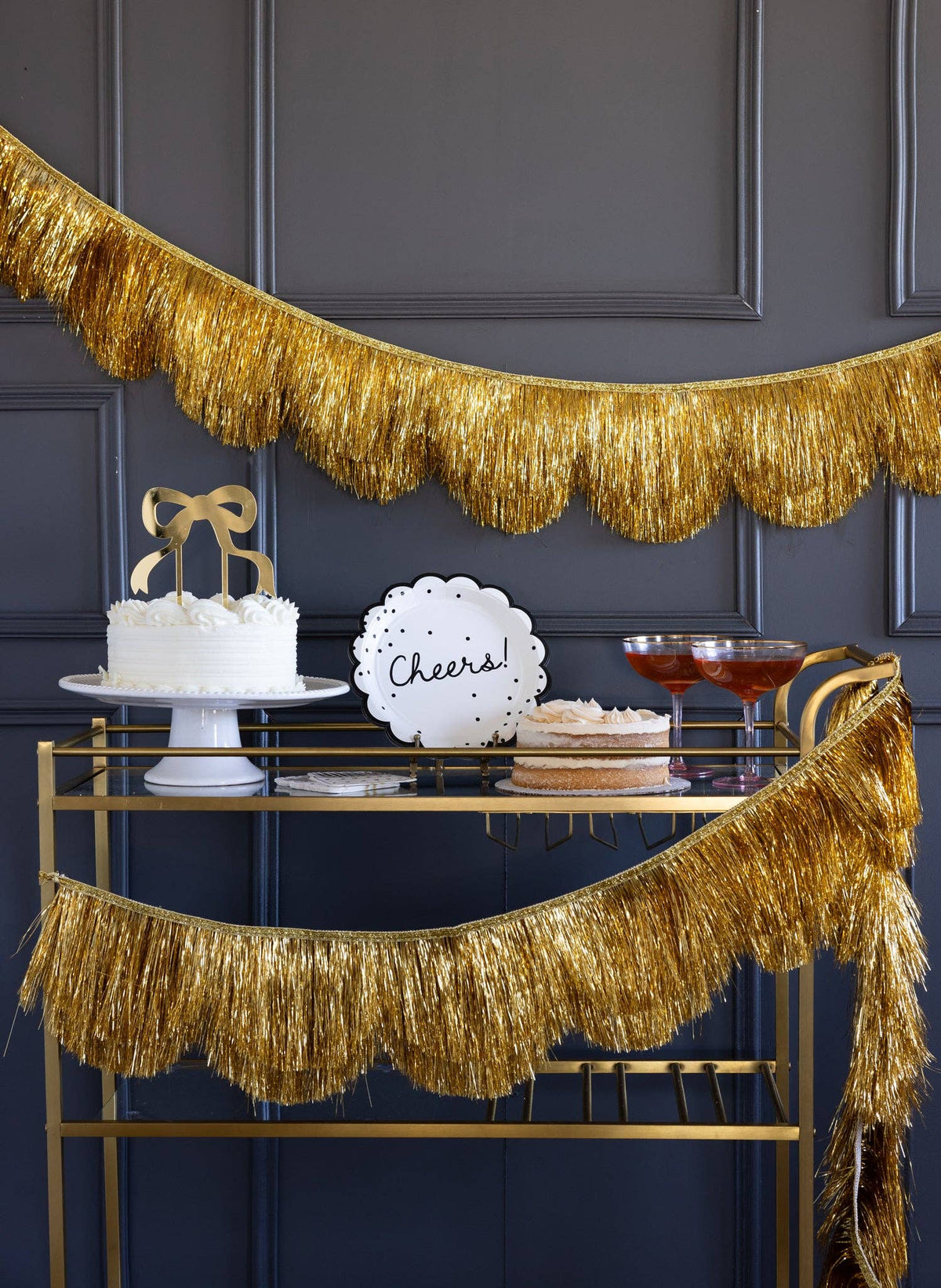 The gold fringe banner styled along a bar cart with a "Cheers" cake topper and cake.