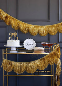 The gold fringe banner styled along a bar cart with a "Cheers" cake topper and cake.