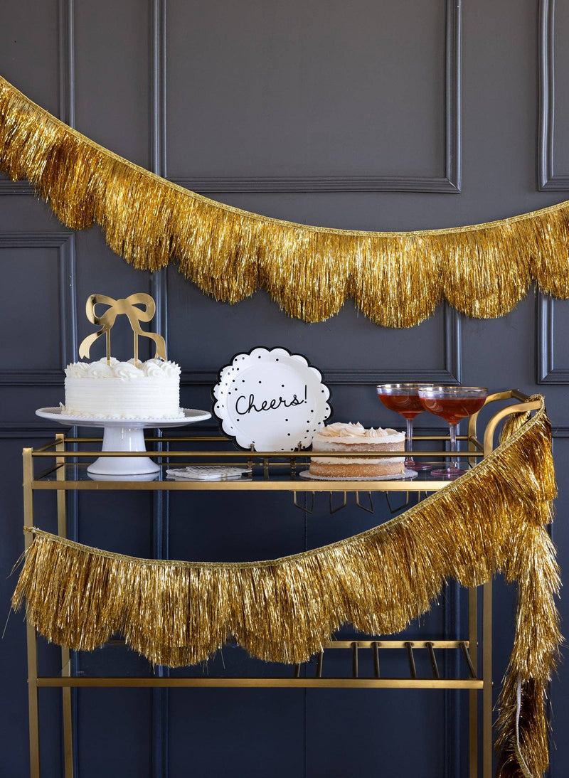 The gold fringe banner styled along a bar cart with a "Cheers" cake topper and cake.