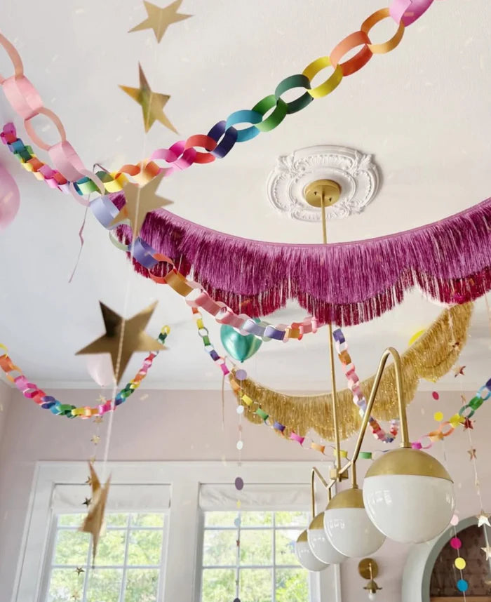 Room styled with colorful paper chain garlands, gold fringe banner, and pink tinsel garland hung from ceiling around chandelier