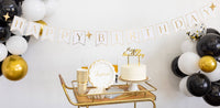 Gold and black birthday party bar cart styled with gold Happy Birthday banner and black white balloon garland, gold cups, Cheers plate, white cake with gold cake topper and champagne flute.