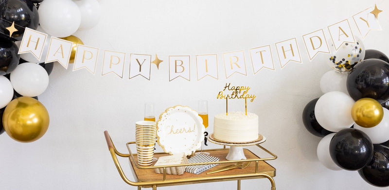 Gold and black birthday party bar cart styled with gold Happy Birthday banner and black white balloon garland, gold cups, Cheers plate, white cake with gold cake topper and champagne flute.