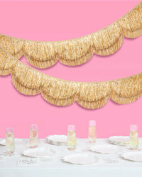 Double-layered gold scalloped fringe banner hanging in two tiers on pink background above styled bridal table with champagne flutes and white plates