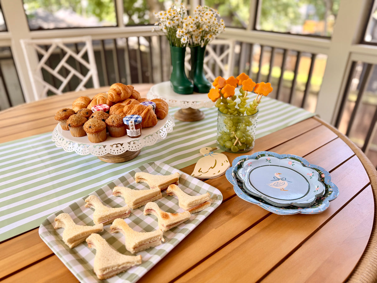 A goose themed table with goose themed paper dinner plates, charger plates, napkins, and sandwiches with a green table runner in the middle.