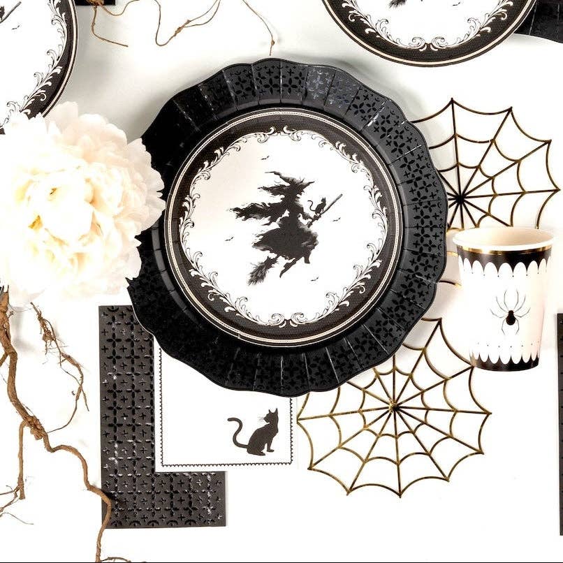 Overhead flat lay of a gothic Halloween tablescape featuring graphic paper spiderwebs, black cat napkins, white florals, and the witch dessert plate.