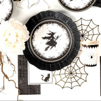 Overhead flat lay of a gothic Halloween tablescape featuring graphic paper spiderwebs, black cat napkins, white florals, and the witch dessert plate.