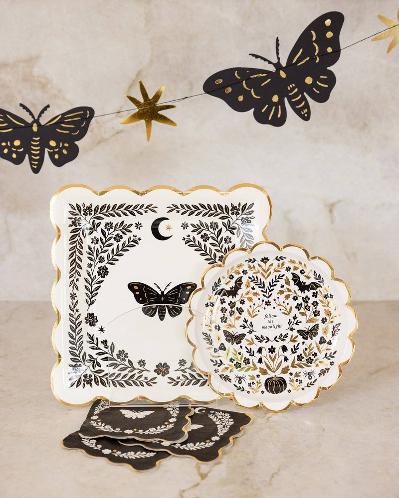 Styled gothic Halloween marble vignette showing the folk-art plate layered with the moth cocktail napkin, black gold-foil moth wall decor, and a gold crescent moon above.