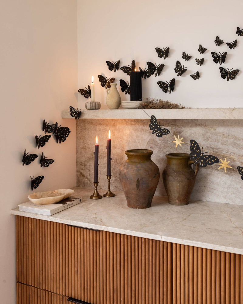 Black gold-foil moths swarming across a marble shelf and wall above a wood-paneled counter with pottery vases and lit candles, moody gothic Halloween styling.