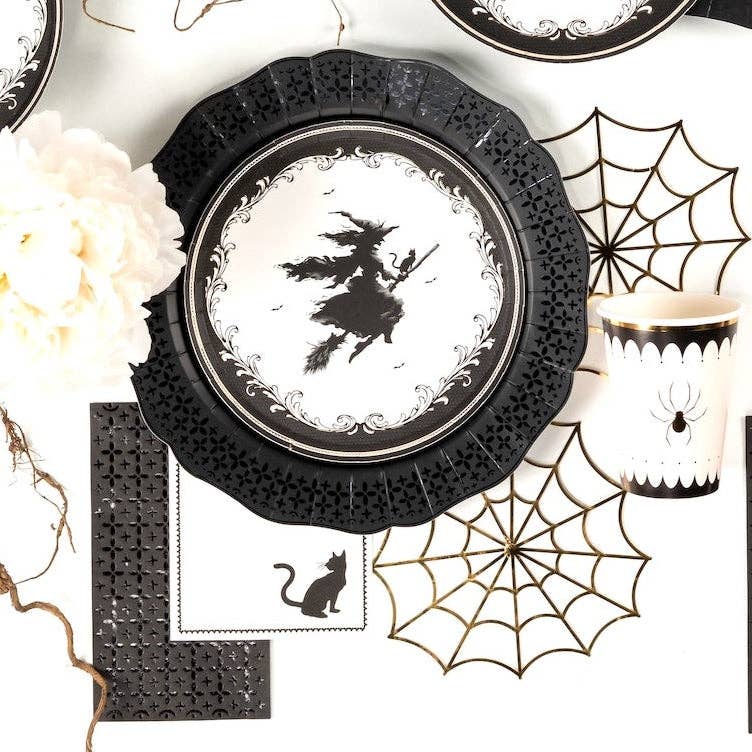 Overhead view of a black lace paper charger, vintage witch plate, and the black cat cocktail napkin styled with a white floral centerpiece.