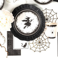 Overhead view of a black lace paper charger, vintage witch plate, and the black cat cocktail napkin styled with a white floral centerpiece.