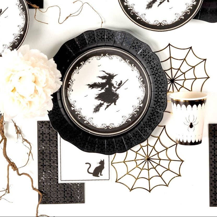 Styled gothic Halloween table overhead — witch silhouette plates on spider web chargers, black-and-white striped runner, gold accents, and black foil guest towels.