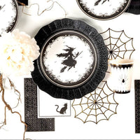 Styled gothic Halloween table overhead — witch silhouette plates on spider web chargers, black-and-white striped runner, gold accents, and black foil guest towels.