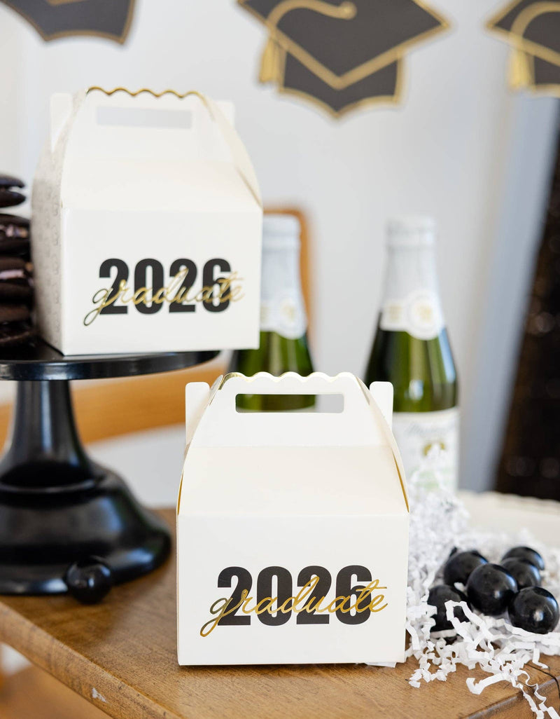 Two white gable boxes with gold foil lettering sitting on a party table next to mini champagne bottles.