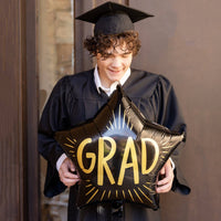 Graduate wearing cap and gown holding black grad star balloon
