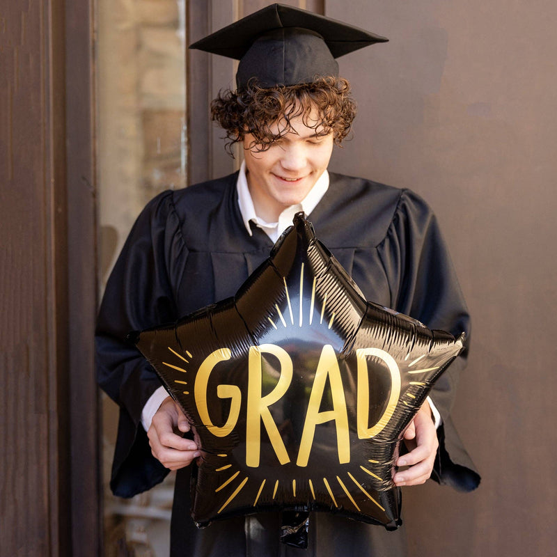 Graduate wearing cap and gown holding black grad star balloon
