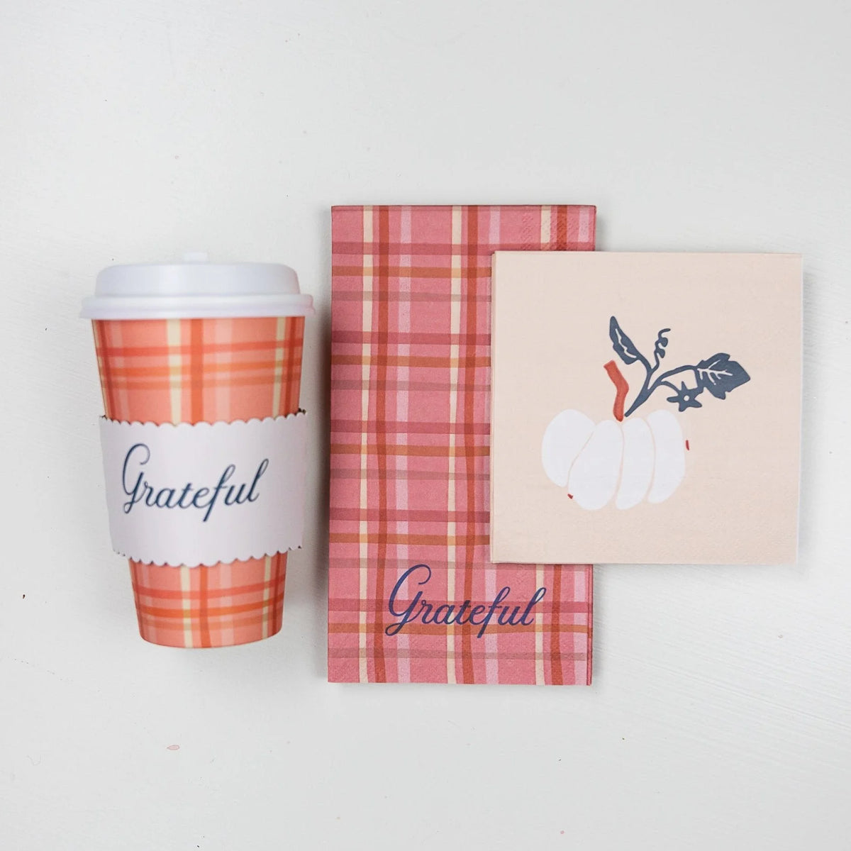 Standing plaid cup with “Grateful” sleeve next to folded plaid guest towel and white-pumpkin napkin on white backdrop.