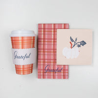 Standing plaid cup with “Grateful” sleeve next to folded plaid guest towel and white-pumpkin napkin on white backdrop.